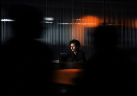 A man at a laptop in a dark room, seen between two blurred figures, an orange light behind him