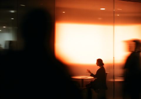 A woman at a table in front of a glowing orange glass wall, blurred people passing in the foreground